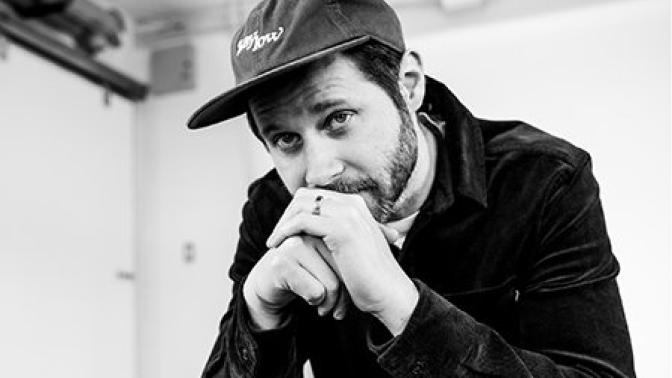 Black and white photo of a man wearing a baseball cap and jacket, resting his chin on his hands and looking thoughtfully into the camera.