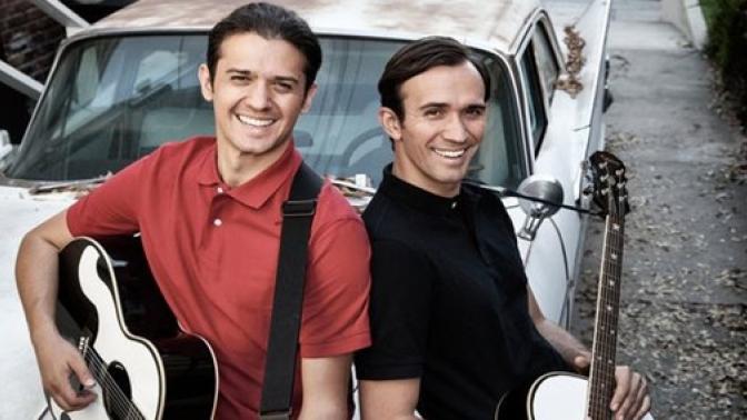 Two smiling men standing back-to-back holding guitars in front of a vintage car.