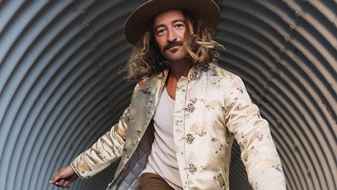 musical artist jesse roper runningthrougha culvert wearing an embroidered cream coat and tan colored hat