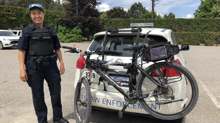 Bylaw officer stands smiling next to a bylaw vehicle with a bike attached to the back.