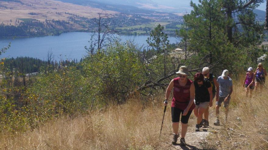 A group of women hike on a trail up a hill with the lake and trees in the distance. 