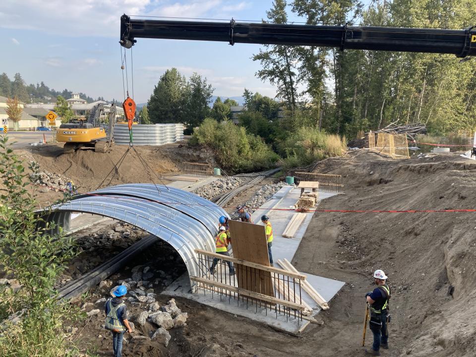 Construction site with workers in safety gear installing a large metal culvert using a crane, surrounded by dirt and trees.