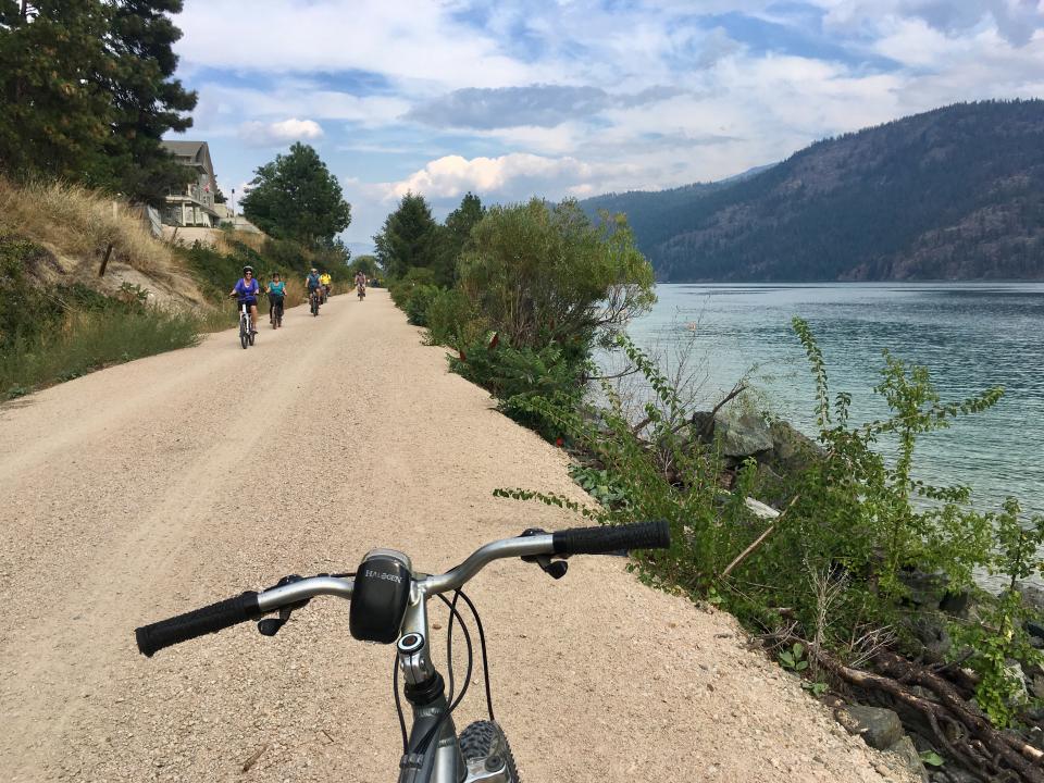 Group of cyclists riding along a gravel path beside a lake with mountainous scenery in the background.