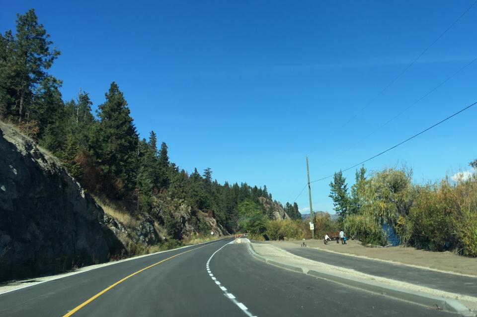 Newly paved road with a bike lane, surrounded by trees and a blue sky.