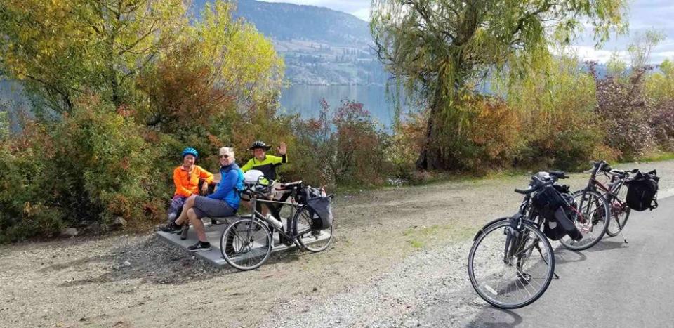 Three cyclists in bright clothing sitting on a bench by a lake, with their bikes parked nearby and mountains in the background.