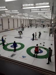 Indoor curling rink with players in action on multiple lanes under bright lights.