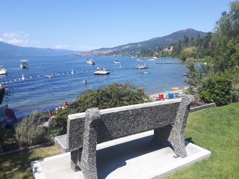 Stone bench overlooking a busy lakeshore with boats, people swimming, and mountains in the background under a clear blue sky.