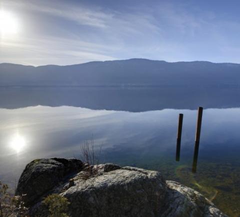 Calm lake with a rocky shoreline, two wooden posts in the water, and mountains in the background under a partly cloudy sky.