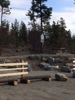 Rustic seating area with wooden benches and picnic tables surrounded by trees and rocks in a forested park.