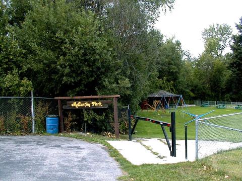 McCarthy Park entrance with a wooden sign, swings, picnic tables, and trees in the background.