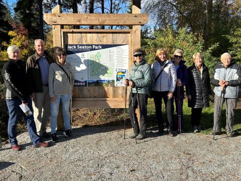 A group of eight people standing in front of a wooden sign for Jack Seaton Park on a sunny day, dressed in outdoor clothing and holding walking sticks.