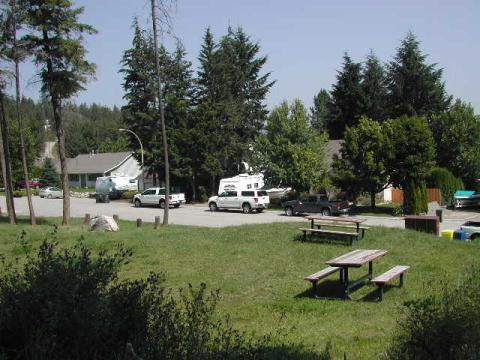 Grassy area with picnic tables and a parking lot with cars and houses in the background, surrounded by trees.