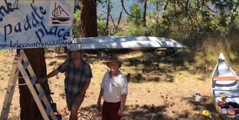 Two men standing near a banner that reads "Paddle Place" with a canoe and a boat on the ground in a wooded area near a lake.