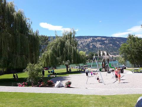 Children playing on swings in a park with large willow trees, picnic tables, and a lake with mountains in the background.