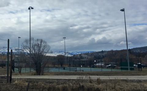 Outdoor tennis courts with tall light poles, bare trees, and snow-covered mountains in the background under a cloudy sky.