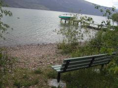 Bench overlooking a rocky lakeshore with a boat docked in the water and mountains in the distance.