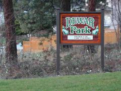 Dewar Park sign with trees and a wooden fence in the background on a grassy area.