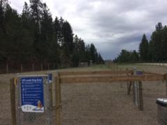 Entrance to an off-leash dog area with a blue sign and fenced gate, surrounded by trees under a cloudy sky.