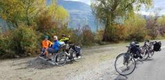 Three cyclists in bright jackets sitting on a bench next to their bikes, with a lake and mountains in the background.