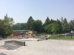 Skate park with ramps in the foreground, playground, trees, and picnic area in the background on a sunny day.