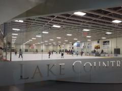 Indoor ice hockey rink with players in action, a referee on the ice, and advertisements along the walls.