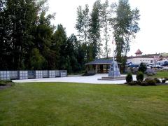 Memorial area with a stone monument, flagpole, and pavilion surrounded by trees and a grassy field.