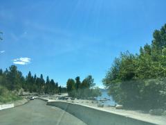 Road along a lakeshore with parked cars, boats in the water, and trees under a clear blue sky.