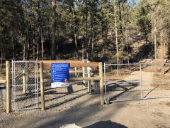 Entrance to an off-leash dog area with a blue sign and a fenced gate, surrounded by trees in a wooded area.