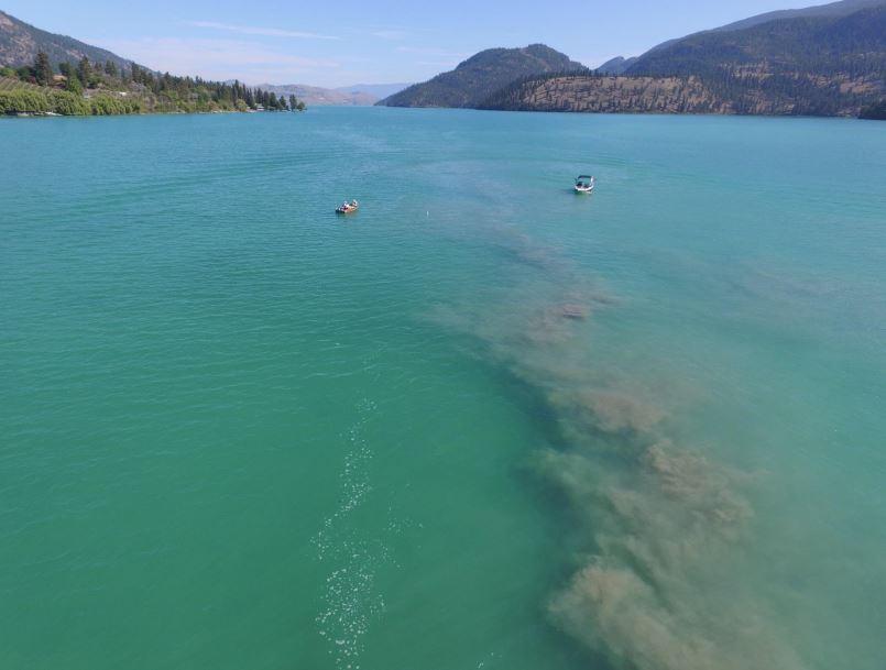 Aerial view of a clear turquoise lake with two small boats, surrounded by forested hills and mountains under a clear blue sky.