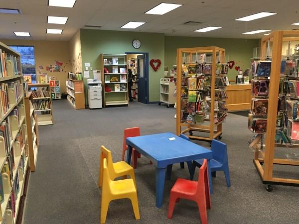 Interior of a library with bookshelves, a small table with colorful chairs, and a counter area.