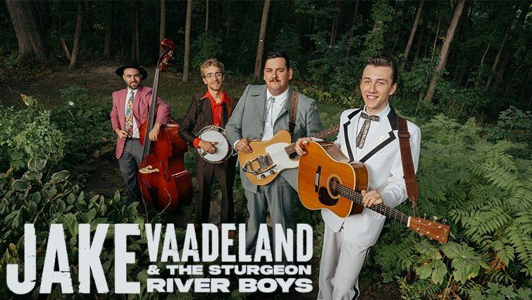 Portrait of Jake Vaadeland holding a guitar with his bandmates holding instruments in a lush green forest. They are all dressed in retro clothing